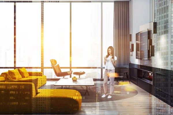 妇女在时尚的客厅内部与白色的墙壁 木地板 黄色沙发 两个黑色扶手椅和电视机挂在壁炉上方 色调图像双曝光 — 图库照片