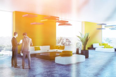 İki adam sarı duvarlar, beton zemin, panoramik pencerelere ve siyah kahve masa ayakta beyaz kanepe oturma odası köşesinde konuşuyor. Tonlu görüntü