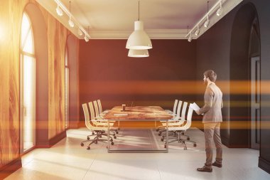 Modern ofis toplantı odası iç siyah duvarlar, kiremitli kat ve beyaz sandalye ile uzun ahşap masa ile ayakta iş adamı. Tonlu görüntü