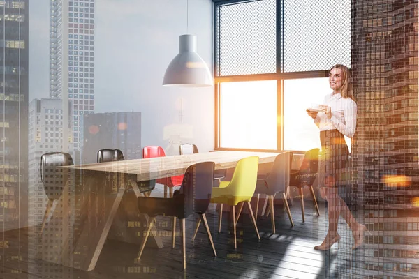 Kadın modern yemek odası ile tuğla ve gri duvarlar, ahşap zemin, geniş pencere ve siyah, kırmızı ve sarı sandalye ile uzun ahşap masa köşesinde. Çift tonlu görüntü pozlaması