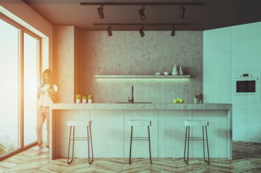 Beton duvarlar, ahşap zemin, beyaz ve beton sayaçları ve dışkı ile beyaz çubuğu ile panoramik mutfakta duran kadın. Çift tonlu görüntü pozlaması