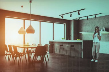 Kadın beton duvarlı panoramik mutfak köşesinde döşeli zemin, gri ve beton sayaçları ve gri bar tabureleri ile. Ahşap masa gri sandalye ile. Çift tonlu görüntü pozlaması