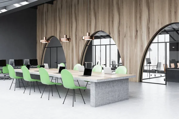 Corner of modern office with green and wooden walls, tiled floor, long wooden and stone table with laptops on it and green chairs. Manager office and meeting room seen through windows. 3d rendering