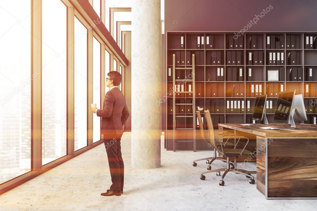 Businessman in interior of company office with gray walls, a column, wooden computer table with brown chairs and bookcase with folders and a ladder. Toned image