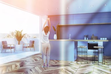 Kadın mavi duvarlar, ahşap zemin, fırın ve lavabo mavi mutfak tezgahı, bar tabureleri ile ve balkon gri koltuklar ile modern mutfak köşesinde. Tonlu görüntü