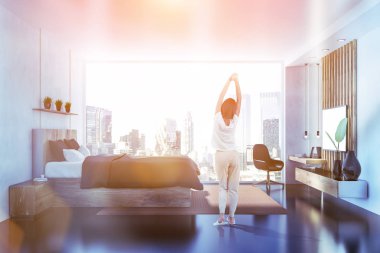 Beyaz duvarları, siyah zemin, panoramik pencere, gri ana yatak ve ev ofis köşe büyük TV ile yatak odası iç yan görünüm ayarlayın. Kadın pijama. Tonlu görüntü