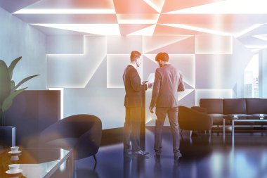 İşadamları modern ofis resepsiyon alanı beyaz geometrik desen duvar, siyah zemin, beyaz ve siyah resepsiyon ve siyah koltuk ve kanepe sehpa yakınındaki köşesinde konuşuyor. Tonlu görüntü