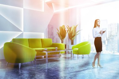 Sarışın işkadını geometrik desen duvarlar, beton zemin ve yeşil kanepe ve koltuklar yakınındaki sehpa ile panoramik ofis bekleme odasında ayakta. Tonlu görüntü