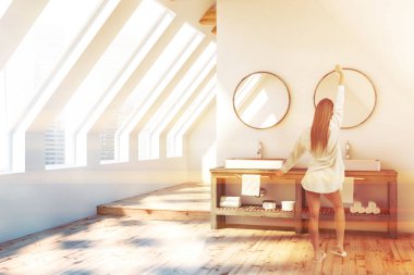 Tavan banyo iç beyaz duvarlar, ahşap zemin ve üstündeki yuvarlak aynalar ile gri tezgah üzerinde beyaz çift lavabo duran kadın. Tonlu görüntü