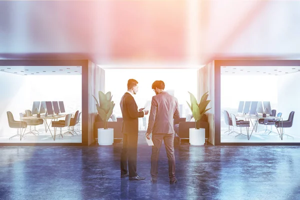 Two white and wooden offices with long wooden computer tables and black chairs in office lobby with two sofas. Businessmen talk. Toned image