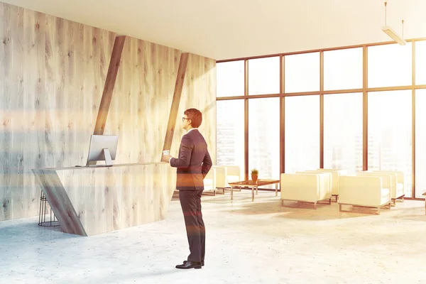 Businessman in corner of office waiting room with wooden walls, concrete floor, wooden reception desk with a computer on it and white armchairs for visitors. Toned image
