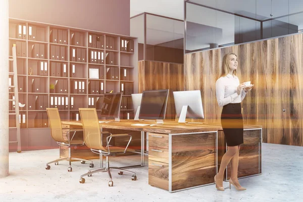 Businesswoman in corner of gray office with wooden computer tables with brown chairs, a bookcase with folders and wooden office doors. Toned image