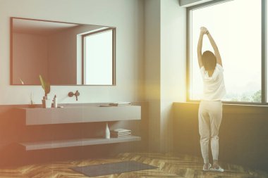 Kadın şık banyo beyaz ve gri duvarlar, ahşap zemin, büyük pencere ve uzun gri lavabo ile yatay ayna üstünde asılı köşesinde. Çift tonlu görüntü pozlaması