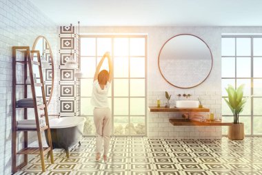 Yan görünüm tuğla duvarlı, kiremitli kat, modern banyo lavabo ahşap raf yuvarlak yuvarlak ayna ve beyaz siyah banyo küvetinde. Havlular için ahşap raflar. Kadın pijama. Tonlu görüntü