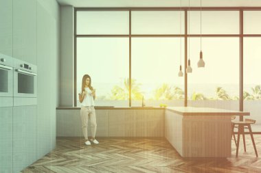 Ahşap zemin, çatı windows, minimalist beyaz mutfak yan görünüm ile beyaz dolap fırınlar ve beyaz bar tabureleri lavabo tezgahı yanındaki ile inşa. Kadın. Çift tonlu görüntü pozlaması