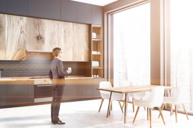 İç siyah tuğla ve gri duvarlar, beton zemin, gri mutfak tezgahı ve ahşap dolapları ile panoramik mutfak ve ahşap masa sandalye ile bir adam. Tonlu görüntü