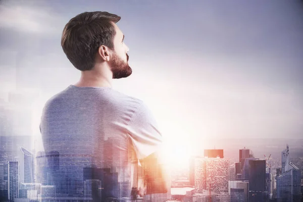 Rear view of thoughtful startup founder wearing t shirt standing over morning city panorama. Toned image double exposure