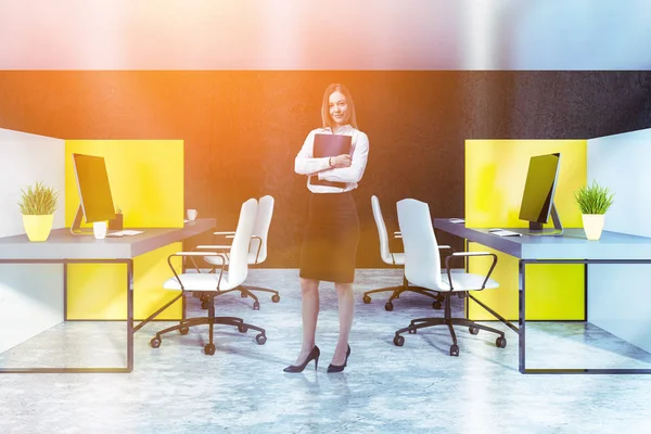 Businesswoman in interior of office with black walls and concrete floor and white and yellow cubicles with gray computer tables and white chairs. Toned image