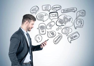 Yan görünümü portresi koyu takım elbise ve onun smartphone ayakta iş hedefi kroki ile gri duvarının yakınında bakarak genç işadamı sakallı