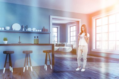 Kadın mavi duvarlar, ahşap zemin, koyu ahşap tabureler olan mavi ve ahşap çubuk, tabak ve oturma odası için kapı ile bir raf ile modern mutfak köşesinde. Tonlu görüntü