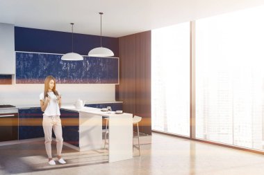 Kadın mavi duvar, taş zemine, koyu mavi mutfak tezgahı ve dolapları ile panoramik mutfak ve dışkı ile beyaz bar köşesinde. Tonlu görüntü