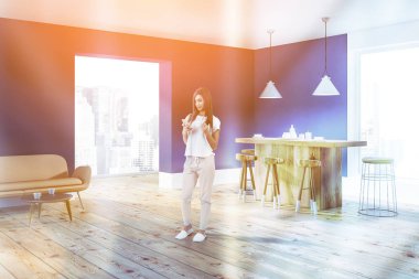 Kadın mavi duvar, ahşap zemin, geniş pencereler ve ahşap bar tabureleri ile minimalist mutfak köşesinde. Kanepe ve bir sehpa. Tonlu görüntü