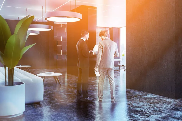 Businessmen talk in corner of office lounge with black walls, concrete floor, long white sofa standing near round coffee tables and office workplace seen in the background. Toned image