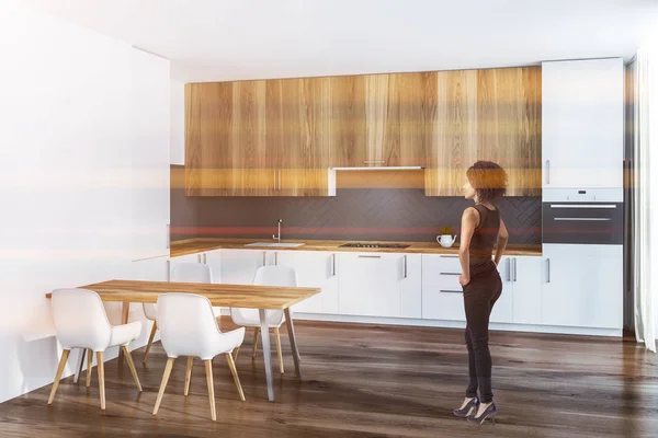 African American woman in corner of kitchen with white and gray walls, wooden floor, white countertops with built in sink and oven and wooden cupboards. Wooden table with white chairs. Toned image