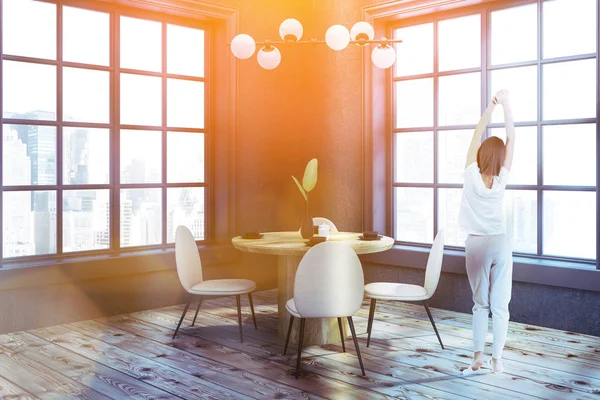 Kadın modern yemek odası ile gri duvarlar, ahşap zemin, köşesinde yuvarlak ahşap masa ve iki büyük pencereler duran bej ve siyah sandalye ile. Tonlu görüntü