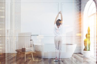 Kadın beyaz duvarlar, ahşap zemin ile minimalist banyo iç ahşap sandalye yanında havlu ile pencere ve beyaz banyo küvetinde kemerli. Çift tonlu görüntü pozlaması