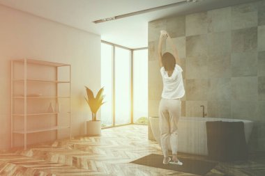 Kadın beyaz ve bej taş duvarlar, ahşap zemin, geniş pencere, gri havlu ile küvet bulunan çatı banyo köşesinde ve şampuan ile beyaz raflar. Çift tonlu görüntü pozlaması