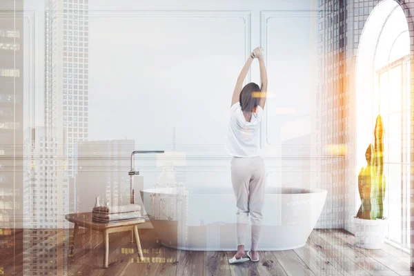 Kadın beyaz duvarlar, ahşap zemin ile minimalist banyo iç ahşap sandalye yanında havlu ile pencere ve beyaz banyo küvetinde kemerli. Çift tonlu görüntü pozlaması