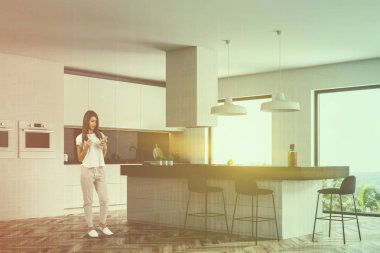 Kadın beyaz mutfak ahşap zemin ile köşesinde, tropikal çatı pencerelerle görüntülemek ve beyaz ve gri ada ile Ocak ve siyah dışkı içinde inşa. Çift tonlu görüntü pozlaması