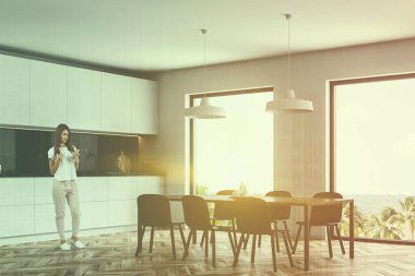 Kadın siyah ve beyaz mermer duvarlar, ahşap zemin, beyaz mutfak tezgahı ve dolapları ile mutfak ve gri yemek masası sandalye ile köşesinde. Çift tonlu görüntü pozlaması