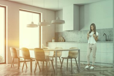 Kadın beyaz duvarlar, ahşap zemin, lavabo ve fırın ile beyaz mutfak tezgahı ve tablo ahşap sandalye ile modern mutfak köşesinde. Çift tonlu görüntü pozlaması