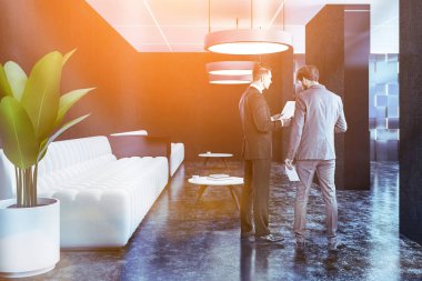 Office lounge iç siyah duvarlı konuşan iki iş adamları, beton zemin ve iki uzun beyaz kanepeler yakın duran sehpa yuvarlak. Tonlu görüntü