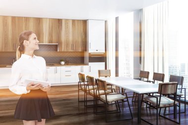 Kadın iç mutfak beyaz ve gri duvarlar, ahşap zemin, beyaz mutfak tezgahı, ahşap dolapları ve uzun beyaz masa sandalye ile. Tonlu görüntü