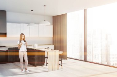 Kadın beyaz duvarlar, ahşap zemin, beyaz ve ahşap mutfak tezgahı ve dolapları ile panoramik mutfak ve dışkı ile ahşap bar köşesinde. Tonlu görüntü