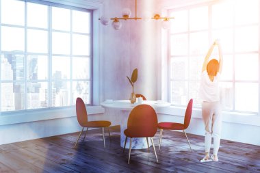 Kadın beyaz duvarlar, ahşap zemin ile modern yemek odası köşesinde yuvarlak beyaz masa ve iki büyük pencereler duran kırmızı ve altın sandalye ile. Tonlu görüntü