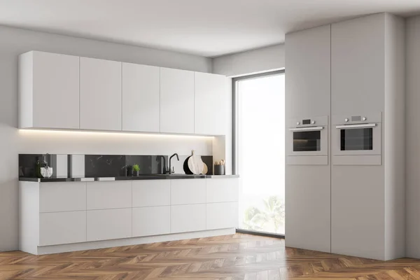 Corner of modern kitchen with white walls, wooden floor, white cupboards, countertops with built in sink and two ovens in the wall. 3d rendering