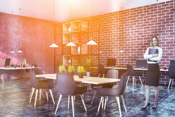 Woman in corner of office with red and brick walls, black floor, long computer table standing near the walls, smaller wooden tables with computers and table with gray chairs in the center. Toned image