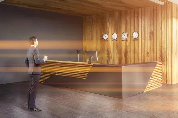 Köşesinde şık gri ve ahşap Resepsiyon Masa bilgisayar ile bu gri ve ahşap duvarlar ile modern ofis saatleri ile ayakta. Kahve ile iş adamı. Tonlu görüntü