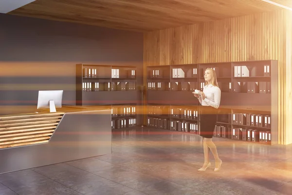 Businesswoman in modern office reception hall with gray and wooden reception desk and black bookcases with folders. Toned image