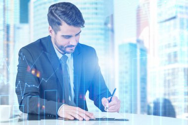 Serious bearded businessman writing in clipboard at office table. Double exposure of blue graphs and cityscape background. Toned image. Stock market concept