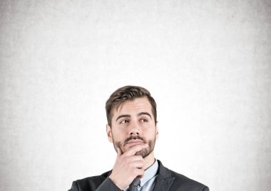 Head and shoulders portrait of young businessman with beard standing near concrete wall and thinking. Mock up