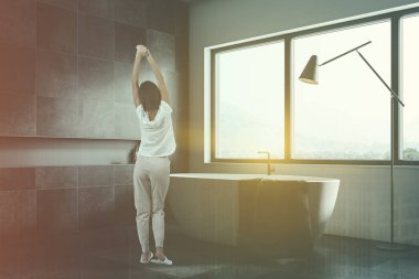 Modern banyo beyaz ve siyah taş duvarlar, siyah seramik zemin, geniş pencere altında beyaz küvet ve şampuan ile raf bir kadın. Çift tonlu görüntü pozlaması