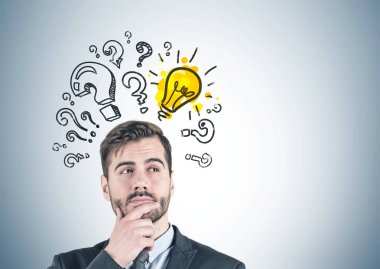 Takım elbise ayakta pek çok soru işaretleri ve ampul üzerinde çizilmiş gri duvarının yakınında sakallı düşünceli genç işadamı portresi. Yukarı alay