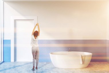 Woman in interior of minimalistic bathroom with white and blue brick walls, concrete floor, white bathtub and white door. Toned image