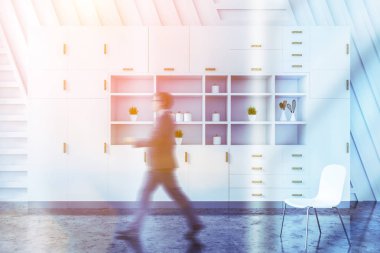 Bardaklı genç işadamı yemek odasındaki beyaz dolabın önünden beton zemin ve beyaz duvarlarla geçiyor. Renkli resim bulanıklaştırıldı