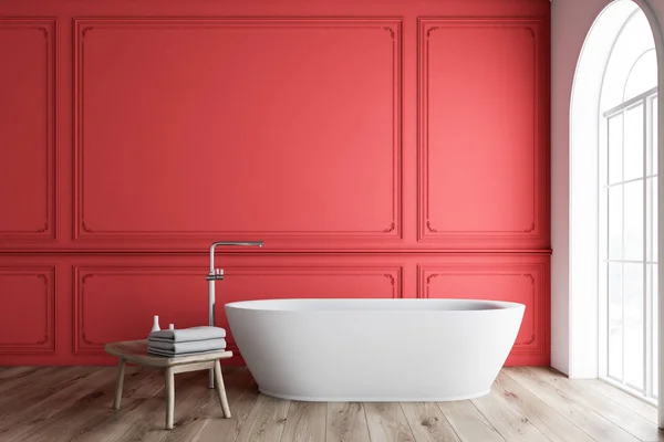 Interior of minimalistic bathroom with red and white walls, wooden floor, arched window and white bathtub with wooden chair with towels next to it. 3d rendering